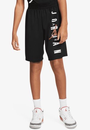 Person trägt schwarze Jordan-Shorts mit vertikalem Logo und weiße Air Jordan Sneaker mit weißen, knielangen Socken vor weißem Hintergrund.