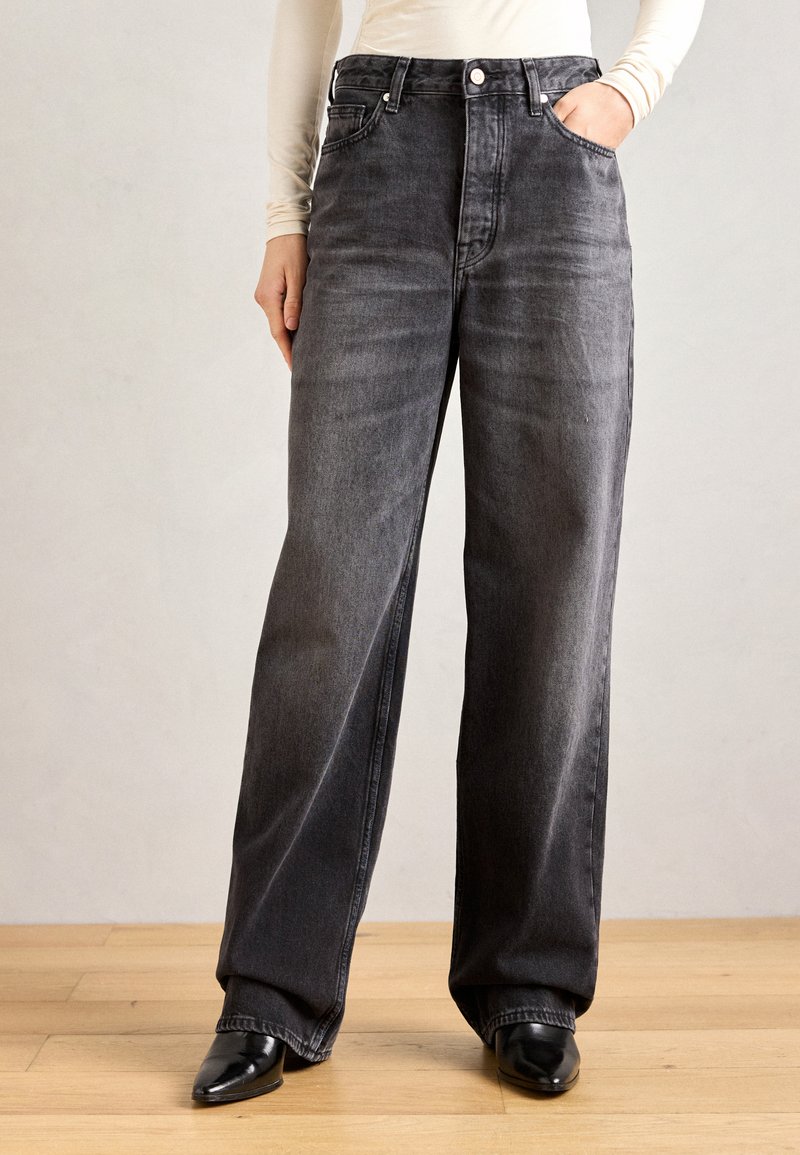 Zwarte denim jeans met wijde pijpen en een hoge taille, voorzakken, en lichte vervaging. Gepaard met zwarte hakken op een houten vloer.
