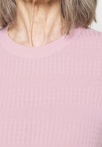 Närbild av en kvinnas hals och hår som bär en strukturerad rosa rundhalsad tröja mot en ljus bakgrund.