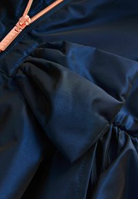 Navyblaue Satinjacke mit einem gerafften Rüschendetail und einem roségoldenen Reißverschluss. Glatte Textur mit Glanz.
