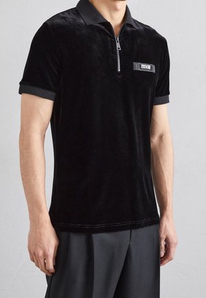 Mann trägt ein schwarzes Polohemd mit kurzen Ärmeln, einem Frontreißverschluss und dem Versace Jeans Couture Logo auf der Brust.