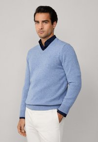 Svetle modrý vlněný svetr s výstřihem do V a žebrovanými manžetami. Nesený přes tmavomodrou košili, v kombinaci s bílými kalhotami.