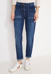 Femme portant un jean jogger en denim bleu retroussé avec de grandes poches avant et des baskets blanches aux accents bordeaux.
