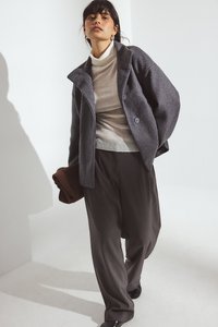 Pelēka vilnas maisījuma jaka ar platām apkaklēm un diviem pogām, valkāta virs gaiša ribota turtlenecka, apvienota ar tumšām brīvā fit stila biksēm.