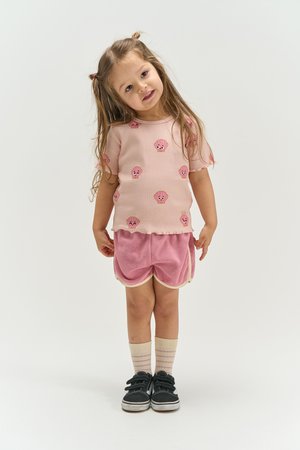 Jong meisje dat met een scheve hoofd staat, draagt een roze shirt met smileygezichten, roze korte broek, gestreepte sokken en zwarte schoenen.