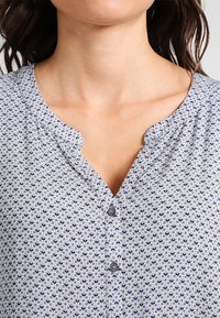 Vaaleansininen pusero, jossa on pieni kukkakuosi, V-pääntie ja neljä harmaata nappia. Kangas on sileä.