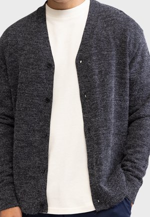 Mand iført en mørkegrå knappet cardigan over en hvid t-shirt med rund hals, med den ene hånd i lommen.