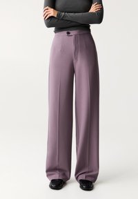 Pantalons larges en tissu lavande doux, avec une taille haute, une fermeture à bouton et des détails plissés à l'avant, associés à des chaussures noires.