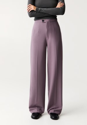 Weite Hosen aus einem weichen Lavendelstoff, mit hohem Bund, Knopfverschluss und eingekreisten Vorderdetails, kombiniert mit schwarzen Schuhen.