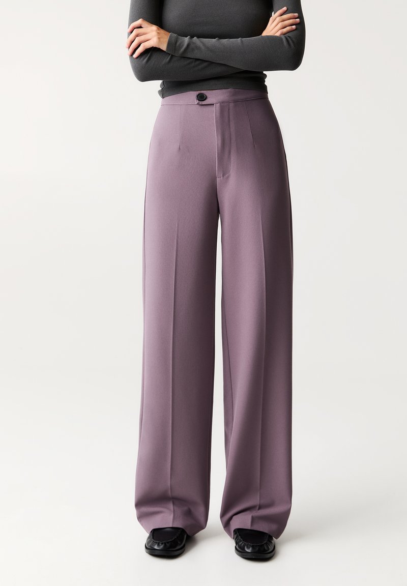 Pantalons larges en tissu lavande doux, avec une taille haute, une fermeture à bouton et des détails plissés à l'avant, associés à des chaussures noires.