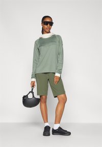 Camicia verde a maniche lunghe con bordi bianchi, abbinata a pantaloni corti olivi. Casco nero tenuto in mano, scarpe sportive nere ai piedi e occhiali da sole scuri indossati.