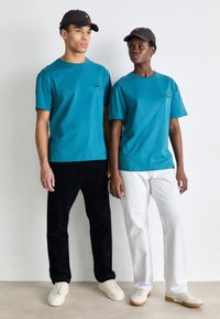 T-shirts en coton teal, coupe confortable, associés à un pantalon en denim noir et blanc. Tous deux portent des casquettes ; l'une est noire avec un logo, l'autre est blanche.