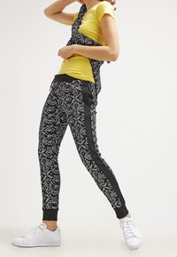 Zwart-witte joggers met een patroontje en zijstrepen, gecombineerd met een gele T-shirt. Model draagt witte sneakers en toont een relaxte pasvorm.