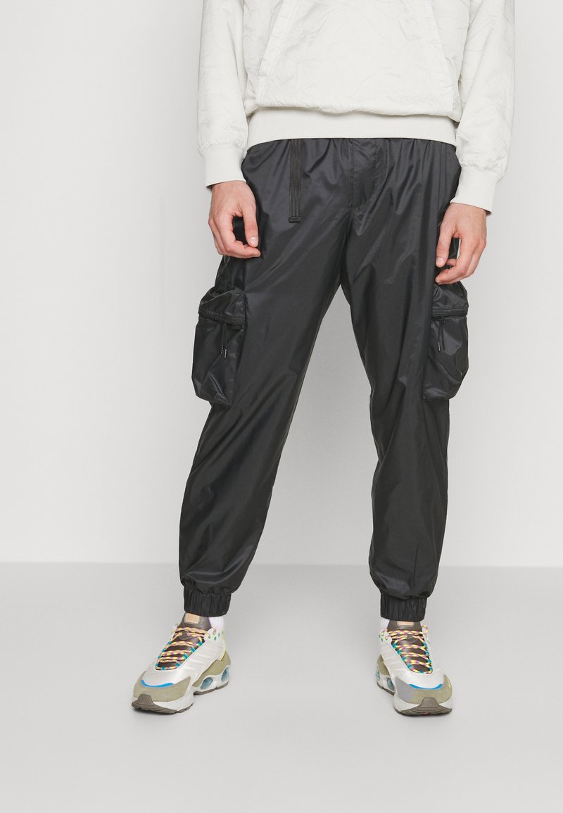 Schwarze Nylon-Cargo-Hosen mit elastischen Bündchen, ausgestattet mit zwei Seitentaschen und einem Kordelzugbund, kombiniert mit mehrfarbigen Sneakers.
