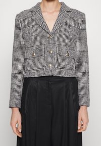 Avskuren, svartvit houndstooth-blazer med spetsig krage, två bröstfickor och guldfärgade knappar. Texturerad tyg, strukturerad passform.