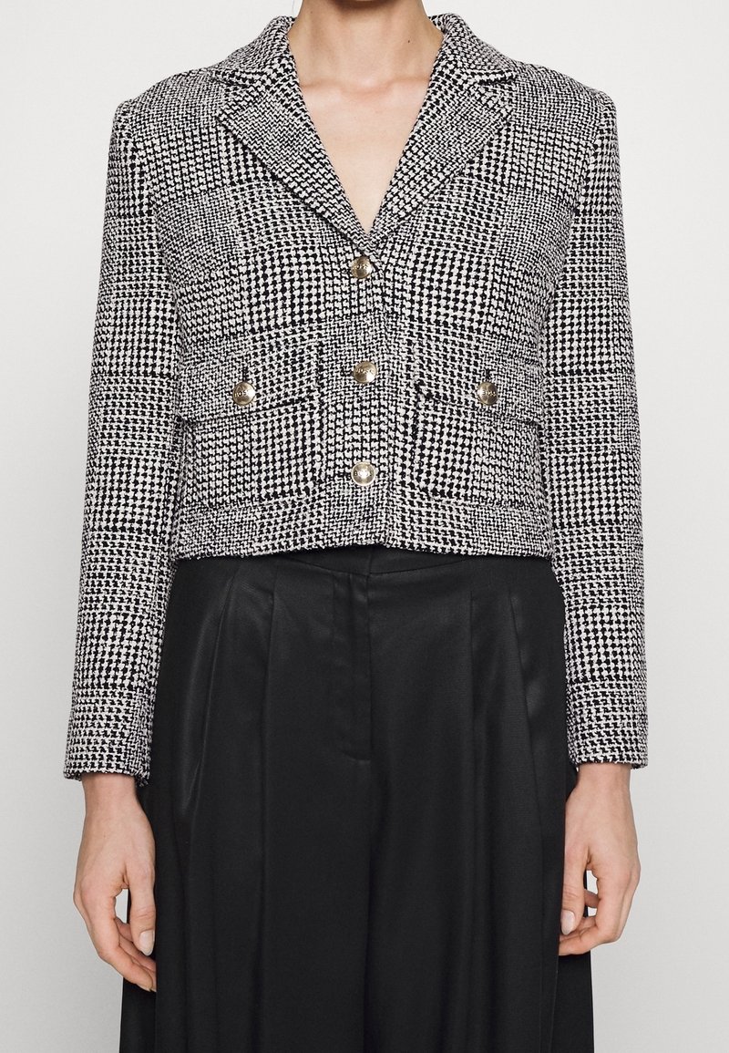 Avskuren, svartvit houndstooth-blazer med spetsig krage, två bröstfickor och guldfärgade knappar. Texturerad tyg, strukturerad passform.