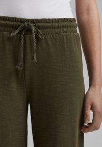 Pantaloni jogger in verde oliva, con texture, dotati di vita elasticizzata e cordino. Mano mostrata che tiene delicatamente la vita.