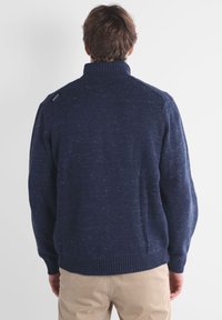 Pull en maille bleu marine avec un col montant, des poignets et un ourlet côtelés, présentant une texture subtile et une marque sur l'épaule gauche.
