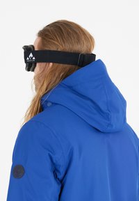 Sininen vedenpitävä takki, jossa on korkea kaulus ja säädettävä huppu. Sisältää mustan laskettelulasihihnan ja logopatchin vasemmassa hihassa.