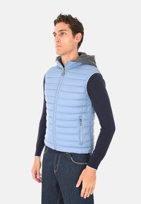 Uomo vestito con un gilet trapuntato senza maniche di colore azzurro chiaro sopra una maglietta a maniche lunghe scura e jeans scuri, in piedi contro uno sfondo semplice.