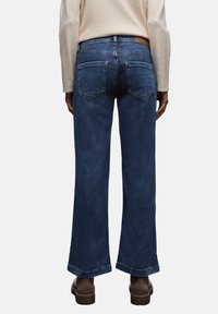 Bredbenta mörkblå denimjeans med bakfickor, som har en läderpatch i midjan och uppvikta benslut. Tyget verkar mjukt med minimal slitage.