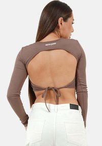 Braun gerippter Langarmtop mit offenem Rücken, mit Bindemöglichkeiten an der Taille und Branding am Kragen. Kombiniert mit weißen Hosen.