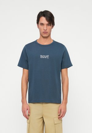 Młody mężczyzna z ciemnymi włosami w niebieskiej koszulce z napisem "Złap świat za ogon" oraz beżowych spodniach cargo, stoi na białym tle.