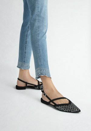 Nogi w niebieskich dżinsach z koralikowymi wykończeniami u dołu oraz czarne baleriny na obcasie typu slingback z wzorem kratki z srebrnych ćwieków, stojące na białej podłodze.