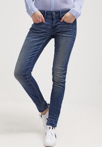 Jeans ajustados en un lavado azul desvanecido, hechos de denim con una textura suave. Presenta cinco bolsillos y un cierre frontal con botón. Combinados con zapatillas blancas.