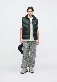 Tânăr în picioare, purtând o vestă matlasată neagră, tricou alb cu imprimeu, pantaloni cu model alb-negru, adidași gri, ținând o șapcă neagră în mâna stângă.