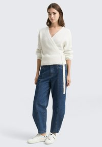 Bela ovita jopica z dolgimi rokavi, iz teksturiranega pletenega materiala, v kombinaciji z ohlapnimi modrimi kavbojkami in belimi supergami.