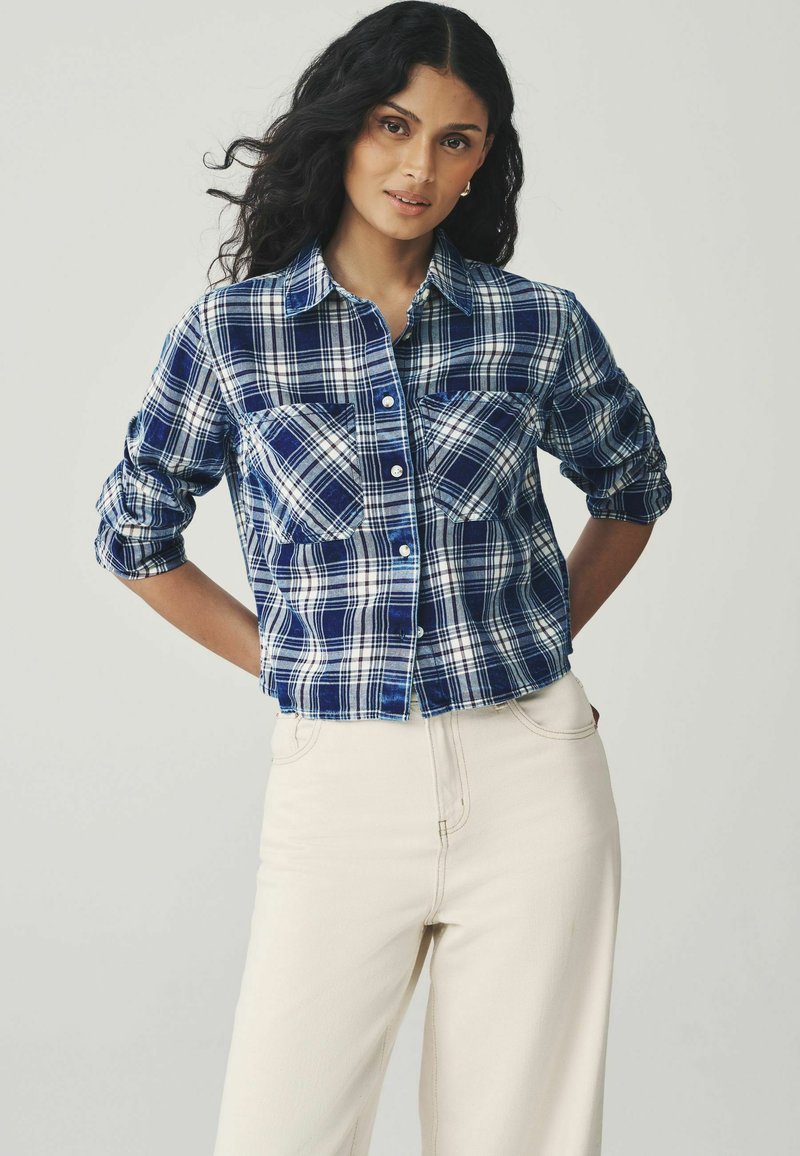 Blue and white plaid shirt with button closure, two chest pockets, and rolled sleeves, made of a lightweight fabric. Paired with beige pants.