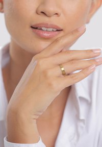 Auksinis metalinis žiedas, dėvimas ant piršto, pasižymintis lygiu, poliruotu paviršiumi. Ranka padėta arti veido, nagai neutralaus atspalvio.