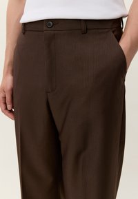 Oseba nosi temno rjave hlače po meri z gumbom na pasu in stranskimi žepi, v kombinaciji z belim zatikanim zgornjim delom.