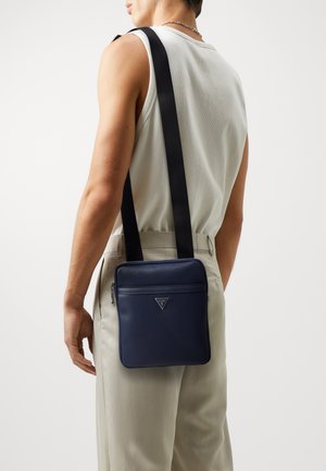 Sac à épaule en cuir texturé bleu marine avec fermeture éclair, sangle réglable et accent logo. Forme carrée, design minimaliste.