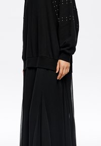 Sudadera negra oversized con detalles de tachuelas en las mangas, combinada con una falda larga negra fluida con aberturas y textura plisada.