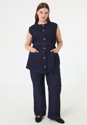 Donna che indossa una tunica senza maniche blu navy con bottoni e cintura e pantaloni larghi coordinati, in piedi con le mani nelle tasche su sfondo bianco.