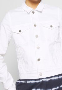 Avkortad vit denimjacka med silverfärgade tryckknappar, två bröstfickor och en platt krage, bärs över en marinblå och vit tie-dye-outfit.