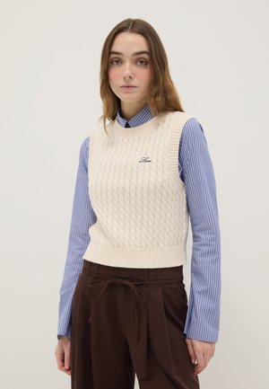 Junge Frau trägt einen cremefarbenen, kabelgestrickten ärmellosen Pullover mit "Love" Stickerei über einem blau gestreiften Hemd und braunen Hosen mit Kordelzug.