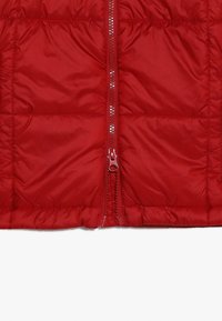 Veste rembourrée rouge avec une fermeture éclair centrée, texture matelassée, matériau léger, et sans logos ni motifs visibles.