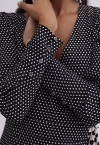 Gros plan d'une personne croisant les bras, portant une chemise noire à manches longues avec des pois blancs et des boutons-pression argentés aux poignets.