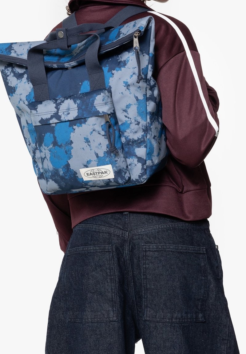 Personne portant un sac à dos Eastpak bleu et gris à motif floral sur une veste bordeaux avec une bande blanche et un pantalon en jean foncé.