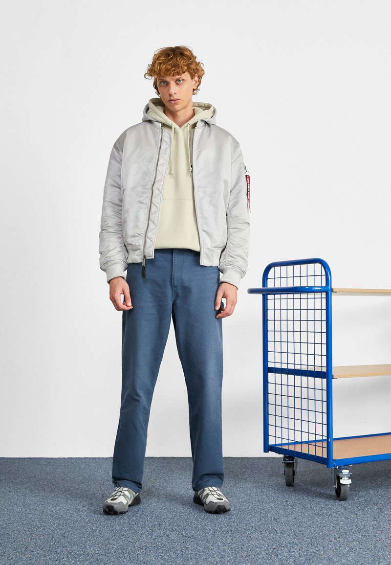 Veste bomber grise claire avec des poignets côtelés sur un hoodie vert clair, associée à un pantalon bleu et des baskets grises. Chariot bleu en arrière-plan.