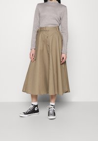 Beżowa spódnica midi z paskiem w talii i detalami w postaci guzików, połączona z szarym, długim rękawem koszulą i czarnymi sneakersami typu high-top.