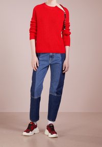 Pull rouge côtelé avec un col asymétrique et un détail zippé. Associé à un jean bleu patchwork et des baskets rouges à carreaux avec des semelles épaisses.