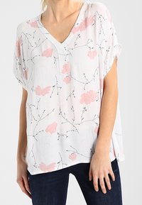 Blouse blanche à encolure en V, ornée de motifs floraux roses et de branches noires. Coupe ample avec manches courtes et tissu texturé.