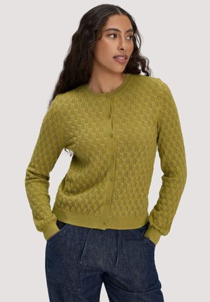 Femme portant un cardigan boutonné vert olive à motif avec manches longues et un pantalon bleu foncé à coupe décontractée, mains dans les poches, regardant vers la droite.