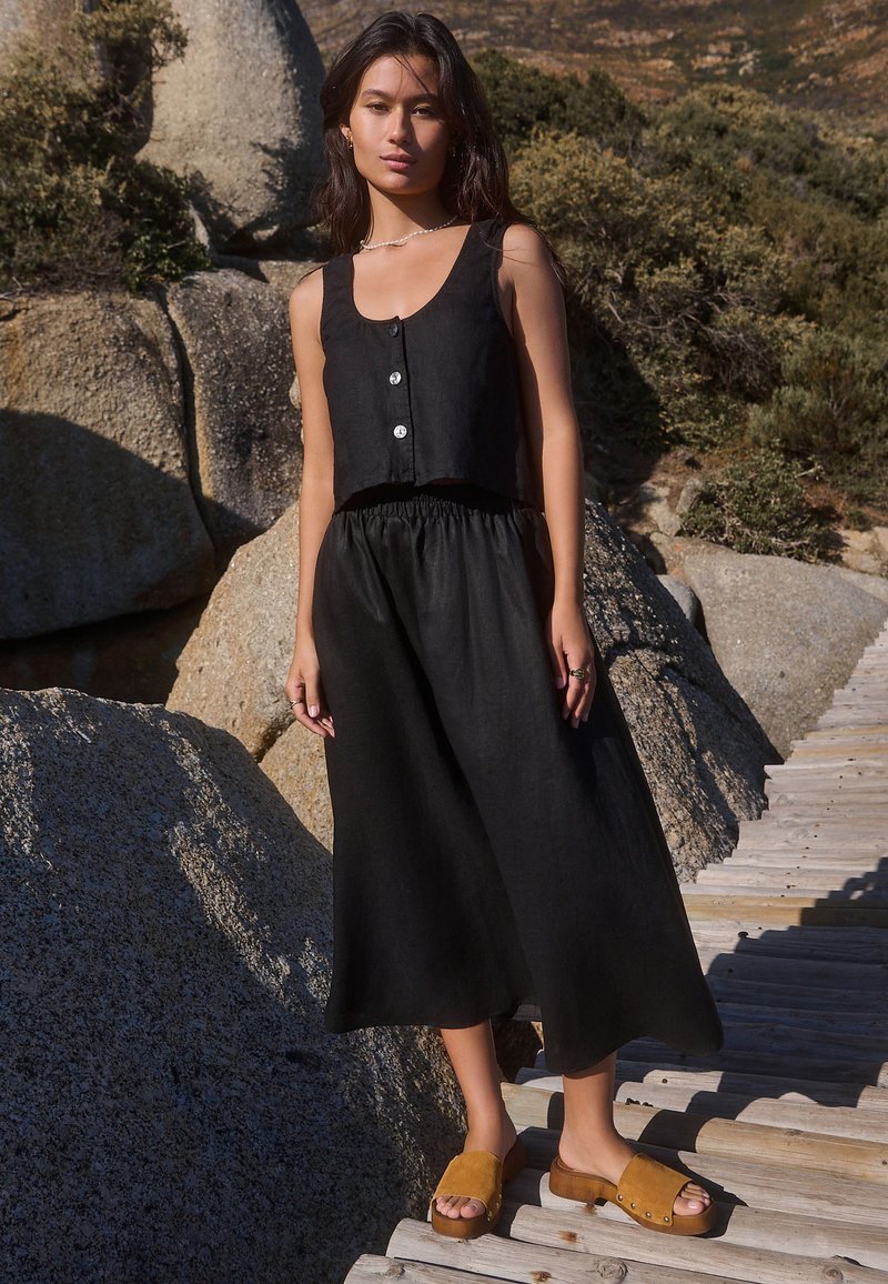 Jeune femme portant un haut noir sans manches boutonné et un pantalon noir à jambes larges, debout sur un chemin en bois parmi de gros rochers.