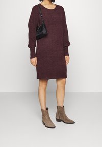 Robe longue à manches longues en tricot de couleur bordeaux avec un col rond, assortie à des bottines beige en daim et un petit sac à bandoulière noir.