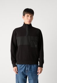 Pullover nero a mezza zip realizzato in tessuto morbido, con taschino sul petto in contrasto e pannelli texturizzati. Abbinato a jeans blu.